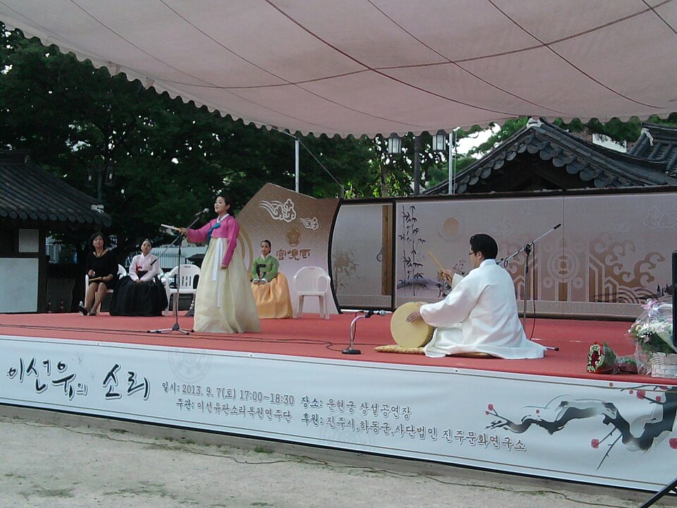 A pansori performance on an outdoor stage: a singer in a pink and yellow hanbok stands, while a drummer in white sits cross-legged with a barrel drum